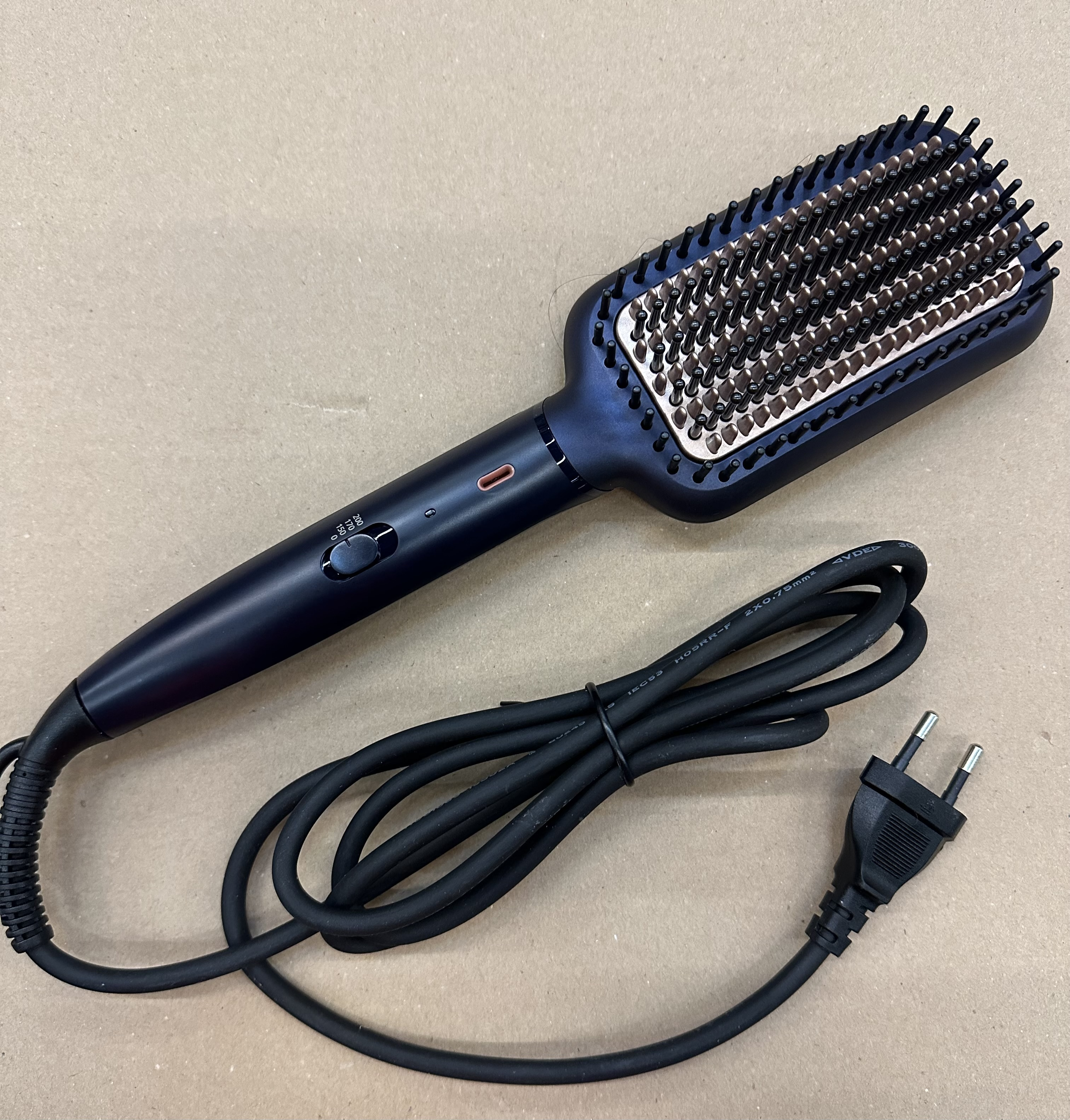 Czarna szczotka do prostowania włosów z kablem i wtyczką, na beżowej powierzchni, gotowa do użycia.