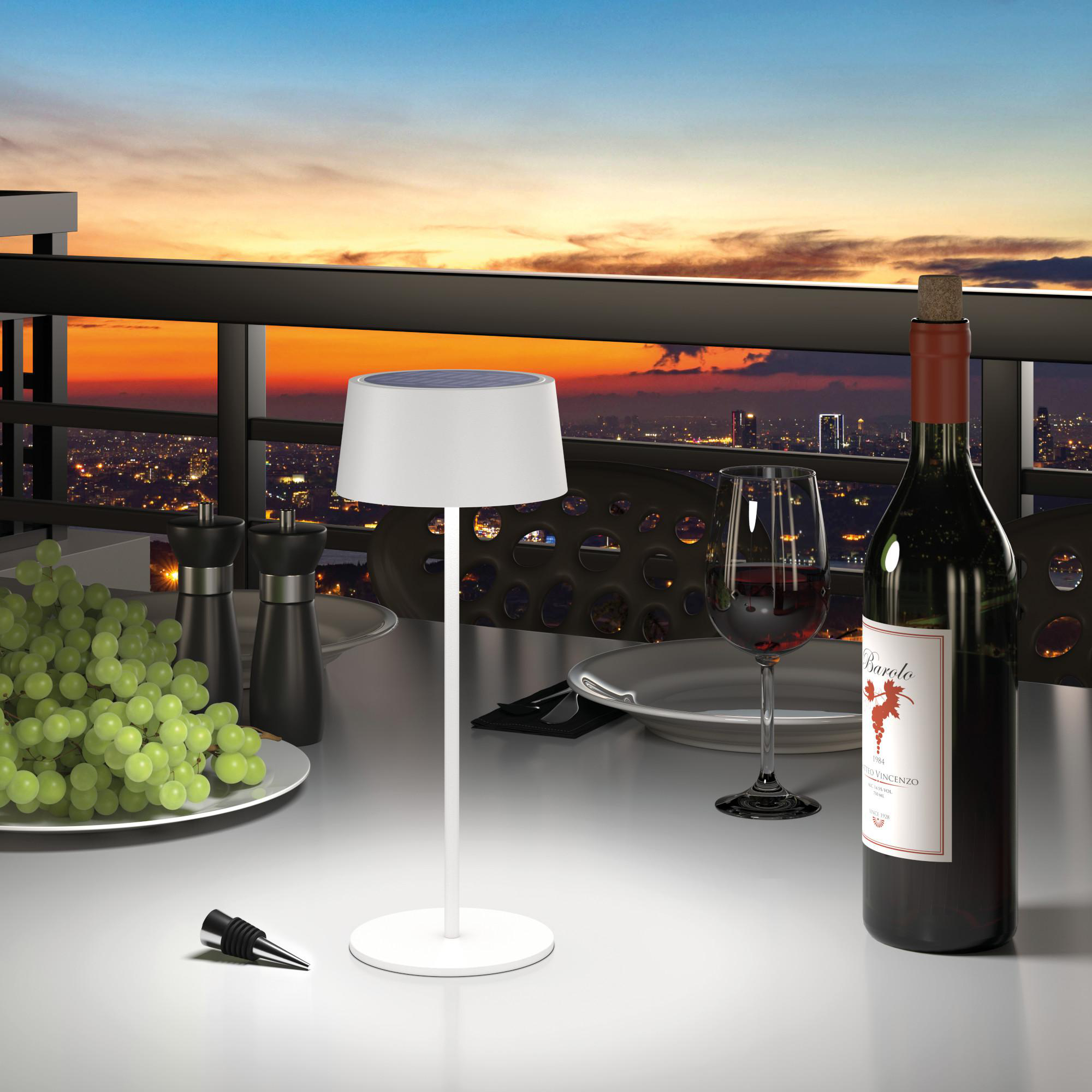 Tisch mit weißer Lampe, Weinflasche, Weinglas, Trauben und Skyline im Hintergrund bei Sonnenuntergang.