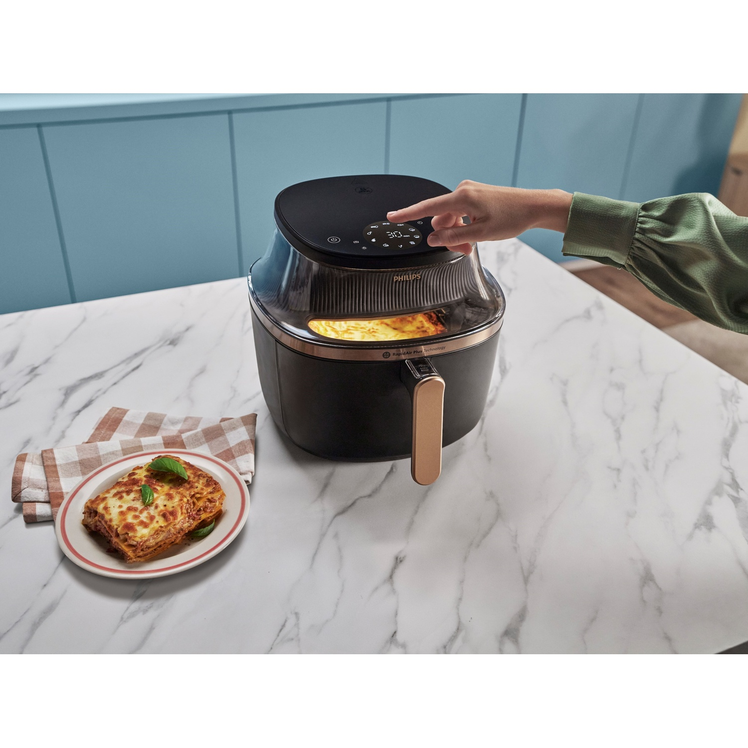 Una friggitrice ad aria con una mano che tocca il display, accanto una lasagna in un piatto.