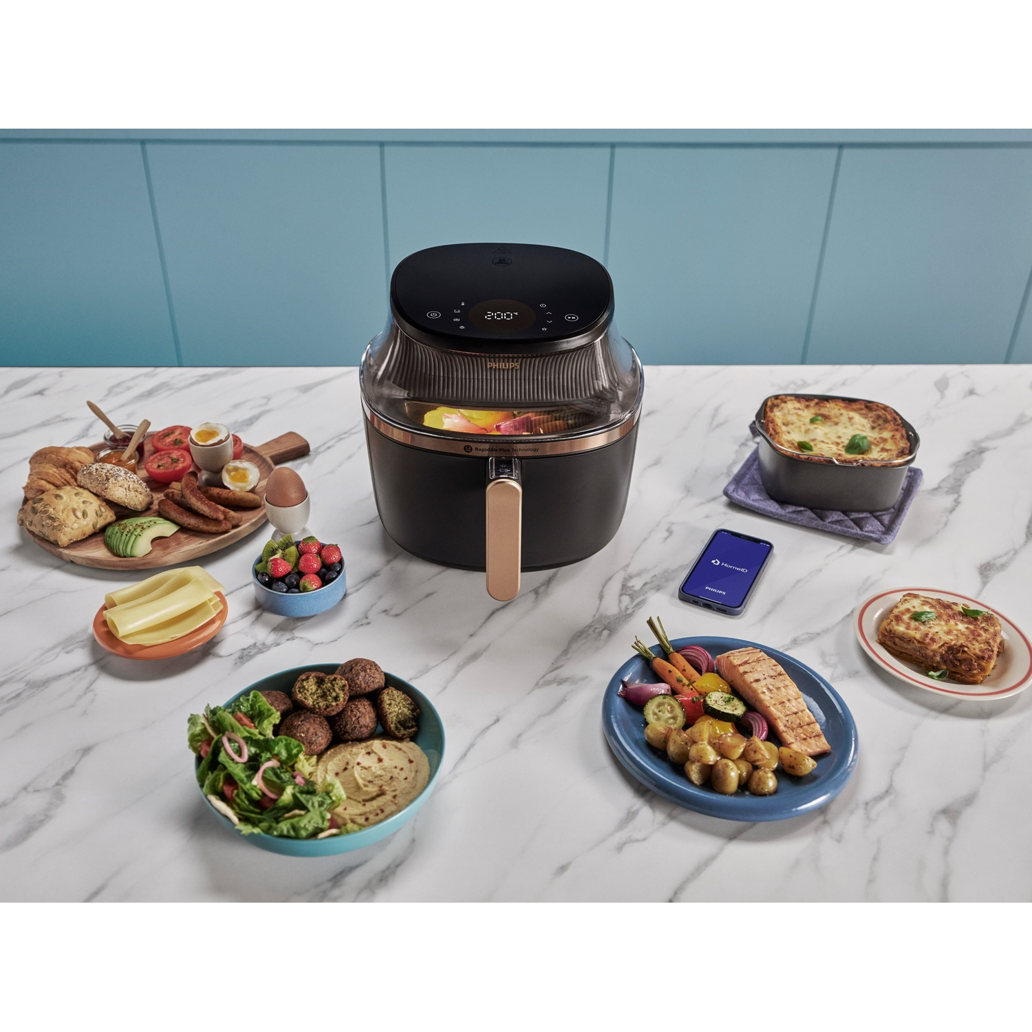 Friggitrice ad aria con cibo vario: verdure, pane, uova, falafel e telefono su superficie bianca.