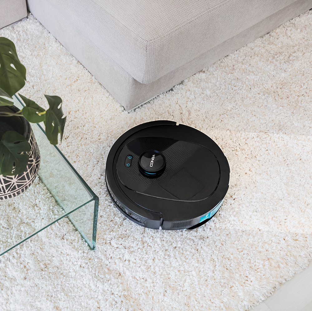 Aspiradora robot negra sobre alfombra blanca y esponjosa junto a un sofá.