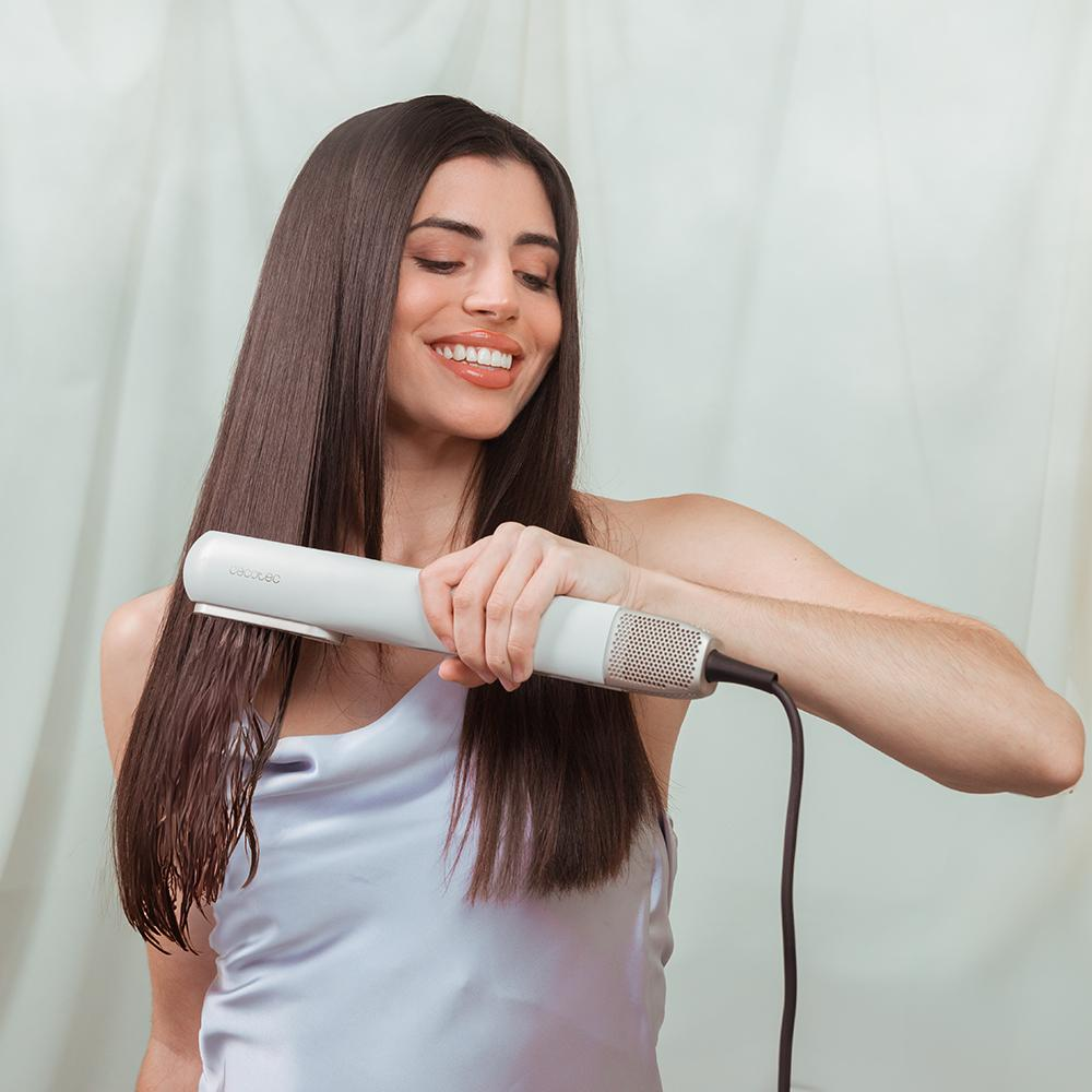 Una mujer sonriendo, usando una plancha de pelo blanca. Su cabello es largo, oscuro y lacio.