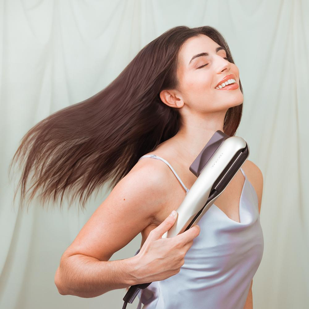 Una mujer con cabello castaño suelto sostiene una plancha de pelo plateada.