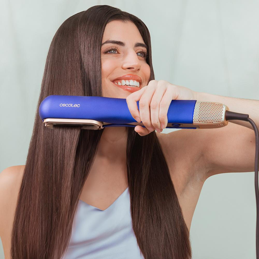 Mujer con cabello largo usando una plancha azul y dorada, sonriendo. El fondo está desenfocado.