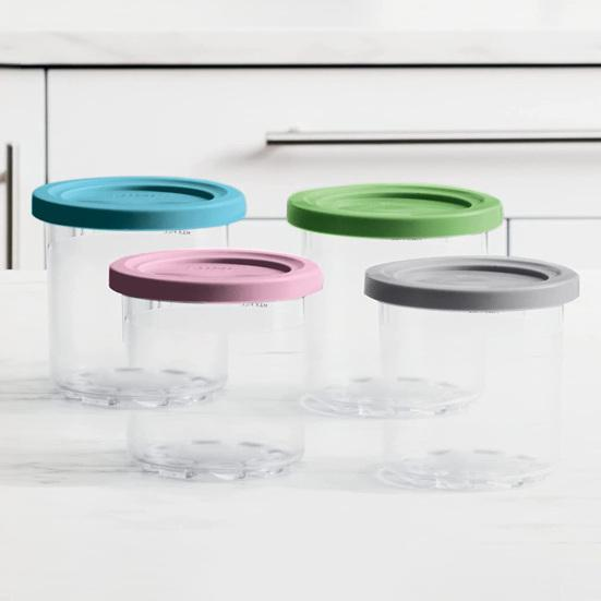 Quatre récipients de stockage transparents avec des couvercles colorés (turquoise, vert, rose, gris) sur une surface blanche.
