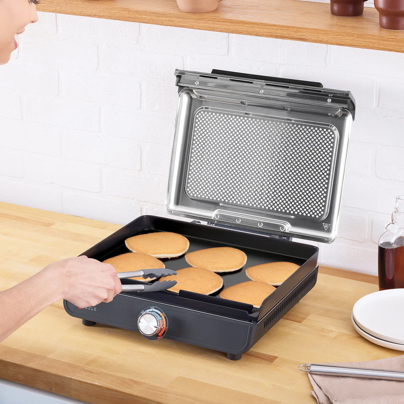 Eine Person benutzt eine Zange, um Pfannkuchen auf einem Grill zu wenden. Der Grill hat eine klare Abdeckung. Holzoberfläche im Vordergrund.