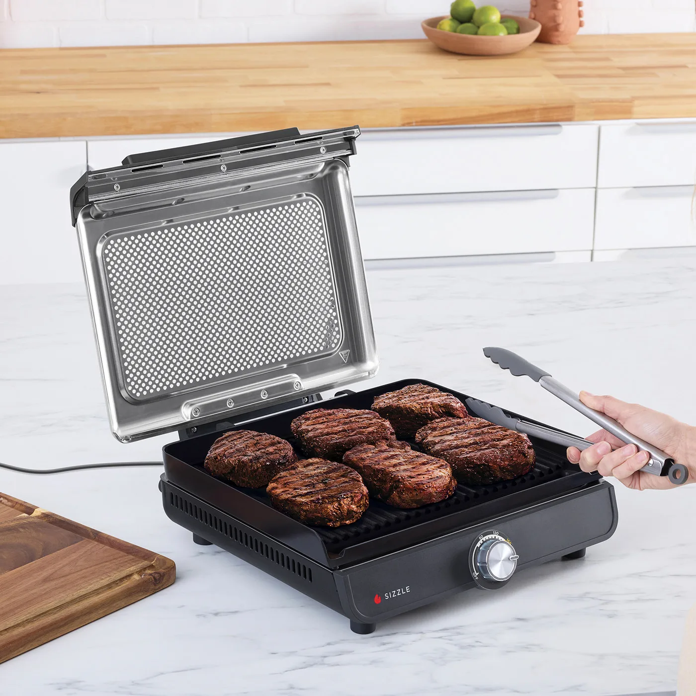 Burger werden auf einem schwarzen Indoor-Grill gegrillt. Eine Hand benutzt eine Zange. Limetten und ein hölzernes Schneidebrett sind in der Nähe. Der Deckel ist offen.