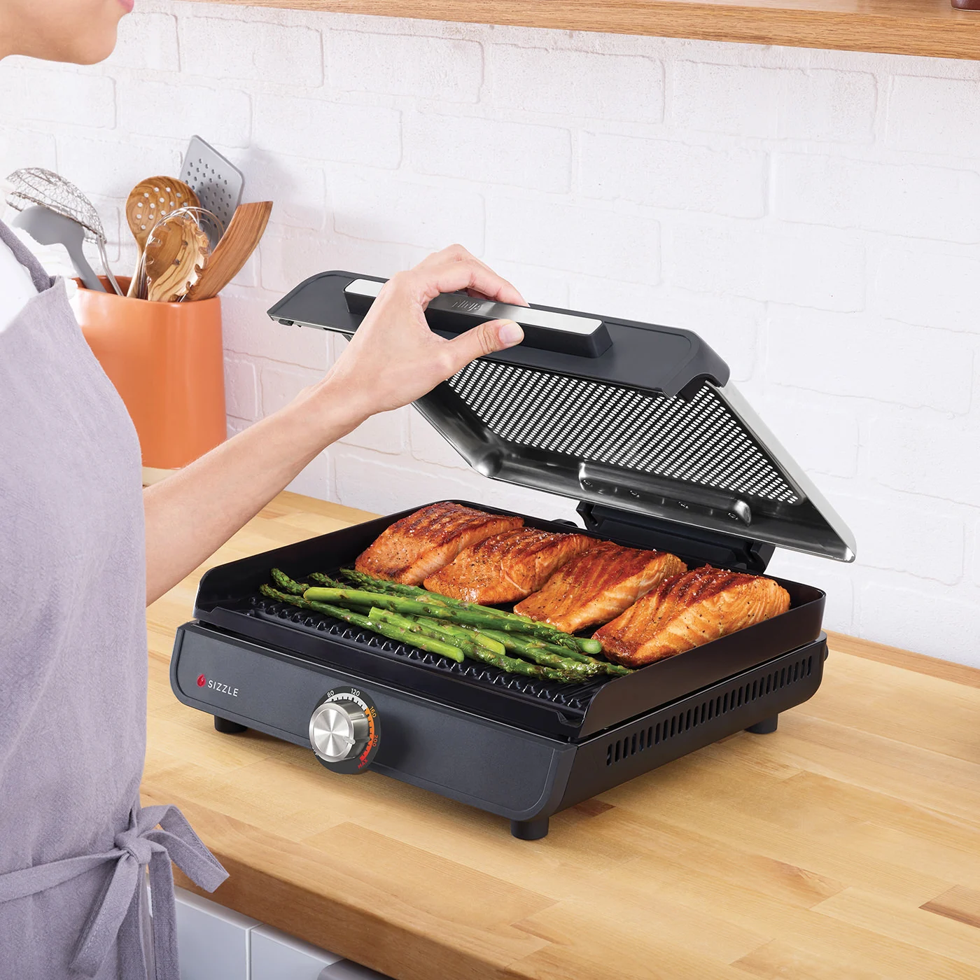 Eine Person öffnet einen Grilldeckel und zeigt Lachsfilets und Spargel. Der Grill steht auf einer Holztheke. Ein Topf mit Utensilien ist sichtbar.