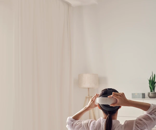 Person trägt VR-Headset, von hinten gesehen. Weiße Wand und Vorhänge.
