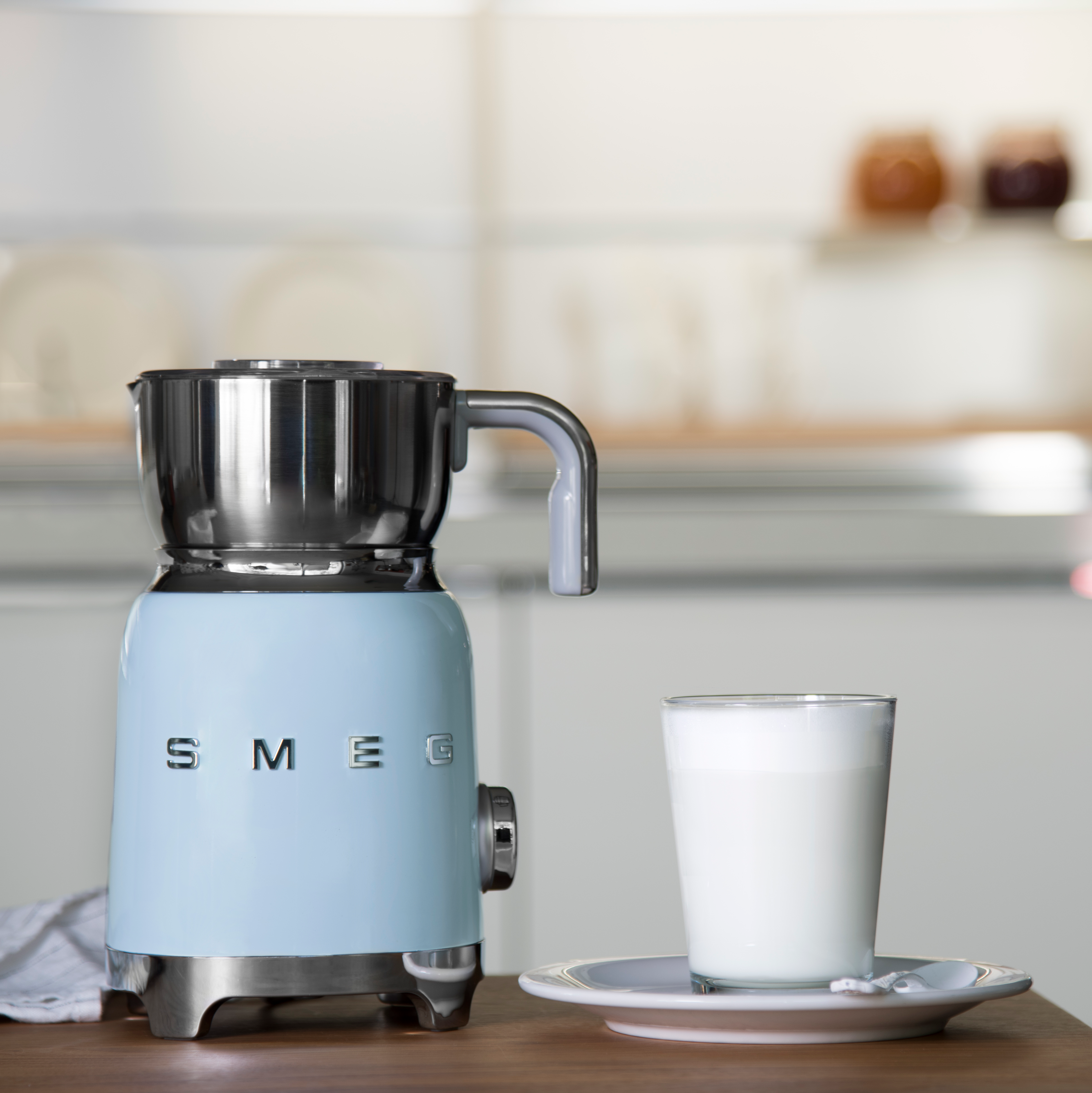 Un mousseur à lait Smeg bleu clair à côté d'un verre de lait sur une surface en bois.