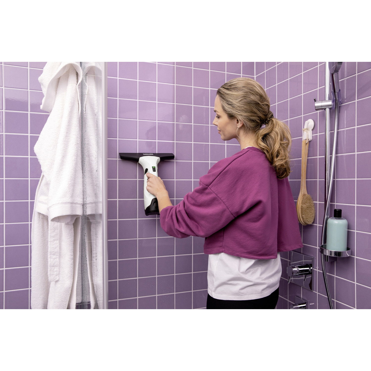 Woman with window cleaner wiping purple tiles in shower, bathrobe hangs.