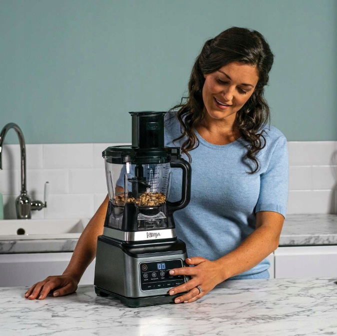 Kobieta w niebieskiej koszuli używa robota kuchennego Ninja, uśmiechając się. Kuchnia z marmurowym blatem.