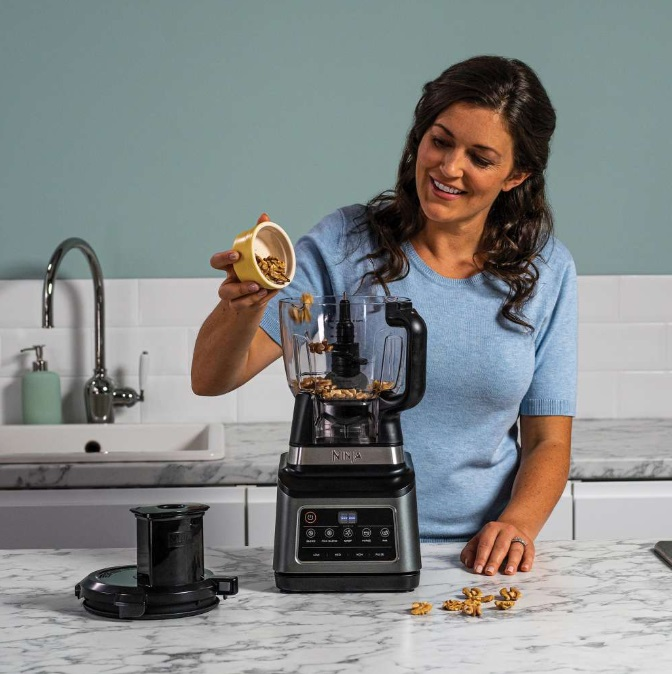 Kobieta dodaje orzechy włoskie do blendera. Urządzenie stoi na blacie kuchennym w pobliżu kranu.