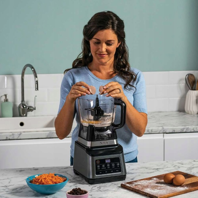 Kobieta wbijająca jajka do robota kuchennego Ninja, uśmiechając się. Kuchnia z marmurowym blatem.