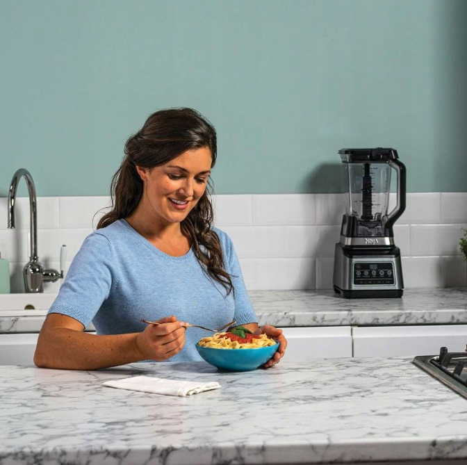 Kobieta w niebieskiej koszuli je makaron z miski, uśmiechając się. Otoczenie kuchni z blenderem.