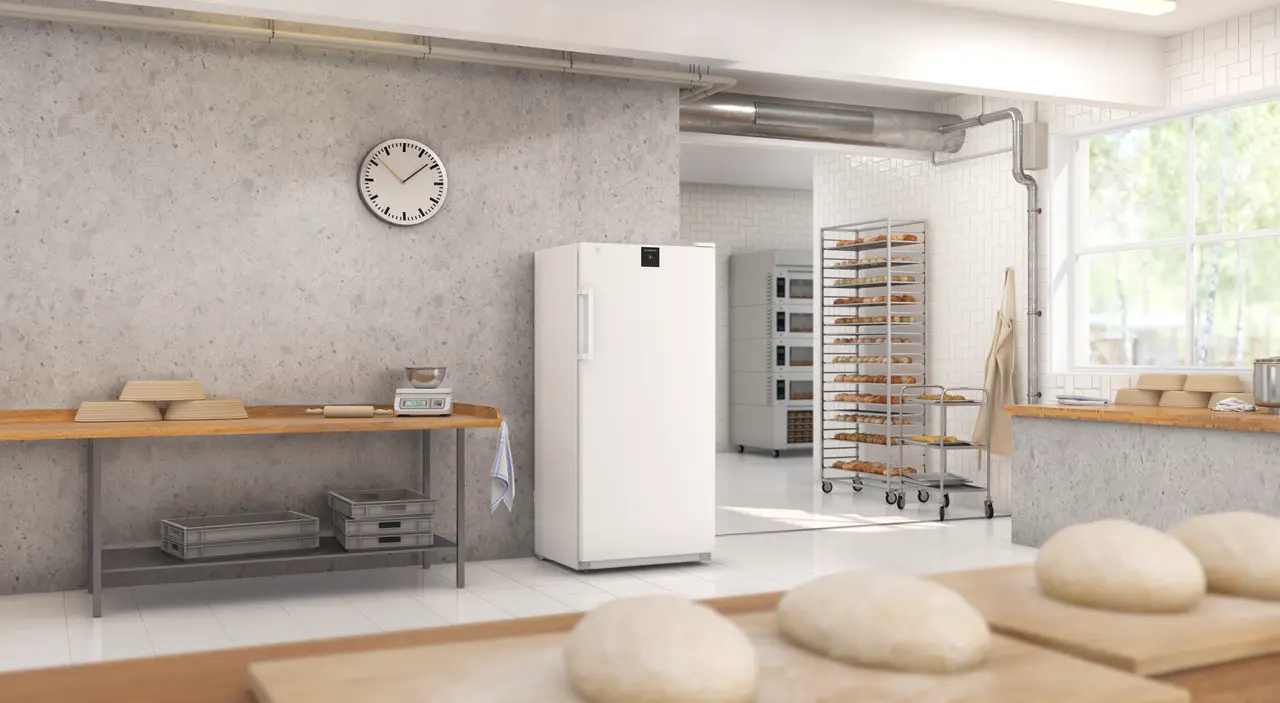 Ein Bäckereiraum. Eine Uhr, ein Kühlschrank und ein Gestell mit Backwaren sind sichtbar.