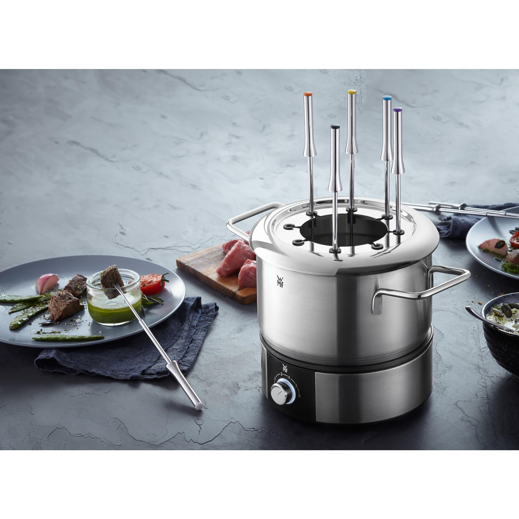 Edelstahl-Fondue-Topf mit Spießen auf einem Tisch. Gemüse und Fleisch liegen daneben.