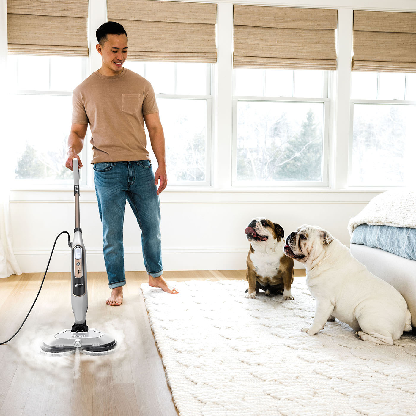 Un homme utilise une vadrouille à vapeur sur un plancher en bois. Deux bouledogues sont assis à proximité sur un tapis blanc.
