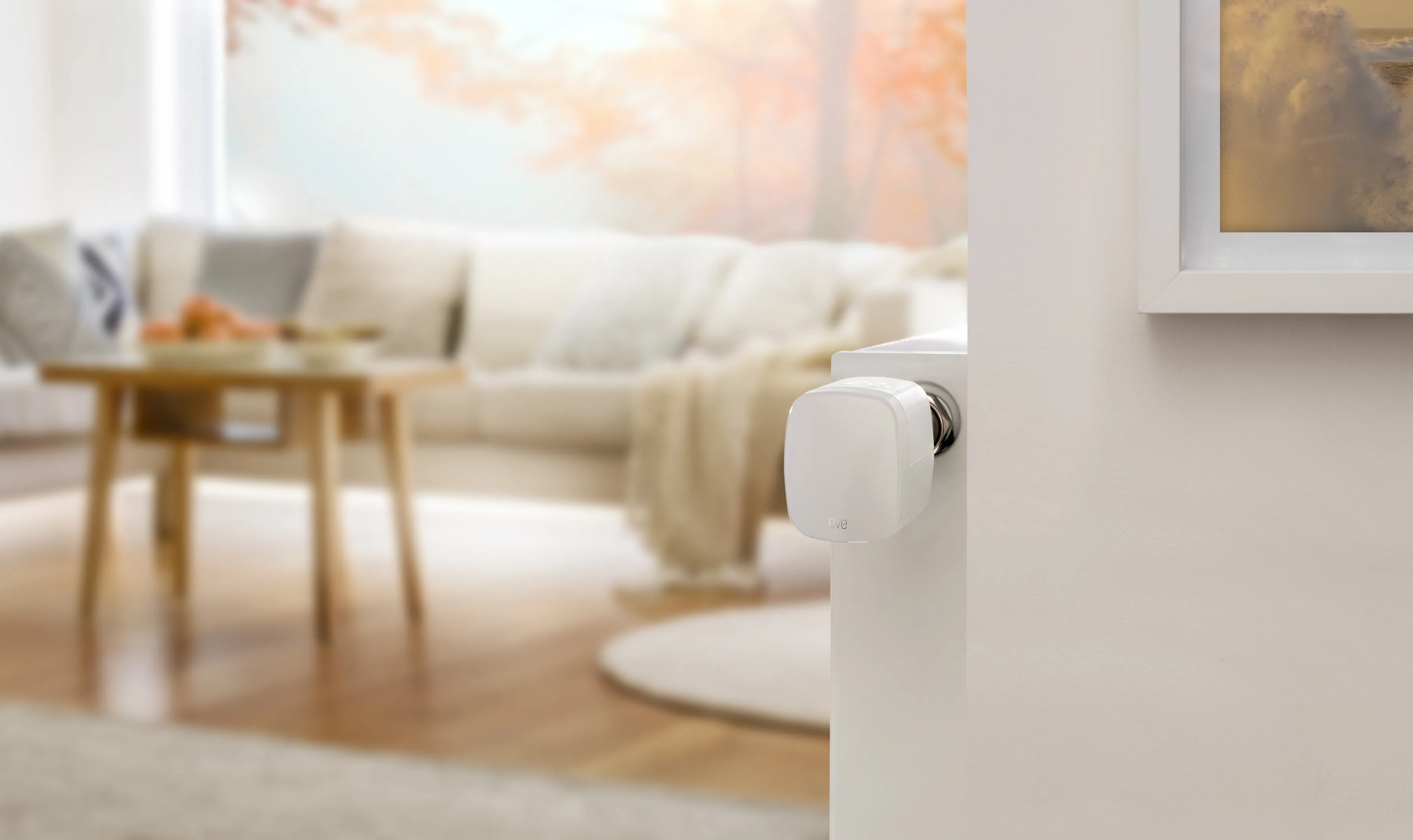 Thermostat intelligent sur un radiateur. Le fond est un salon avec une table, un canapé et une peinture.