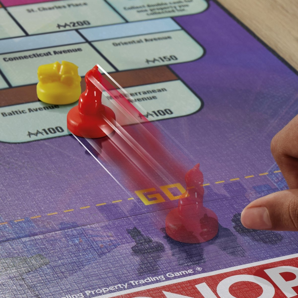 Monopoly game in progress. Red token is moving across the board. A hand is pointing to the Go space.