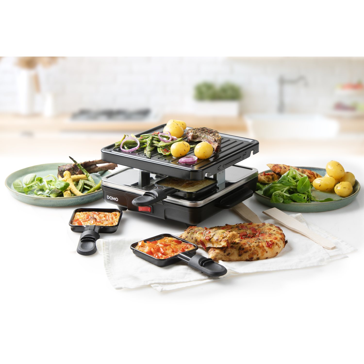 Raclette grill with food, plates, and spatulas. Kitchen setting with white background. Brand name DOMO.