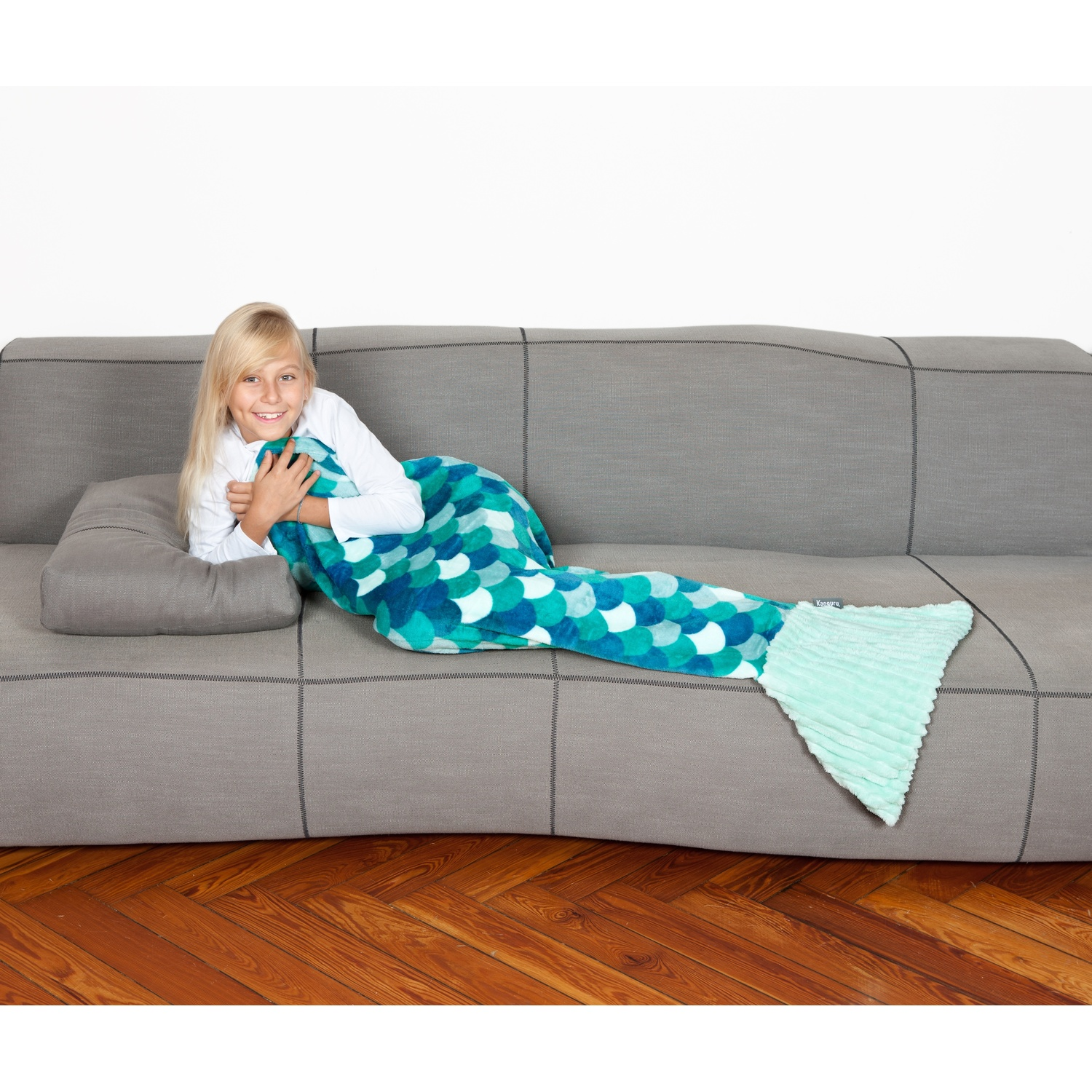Una ragazza è sdraiata su un divano grigio, avvolta in una coperta a coda di sirena blu e verde. Sorride.