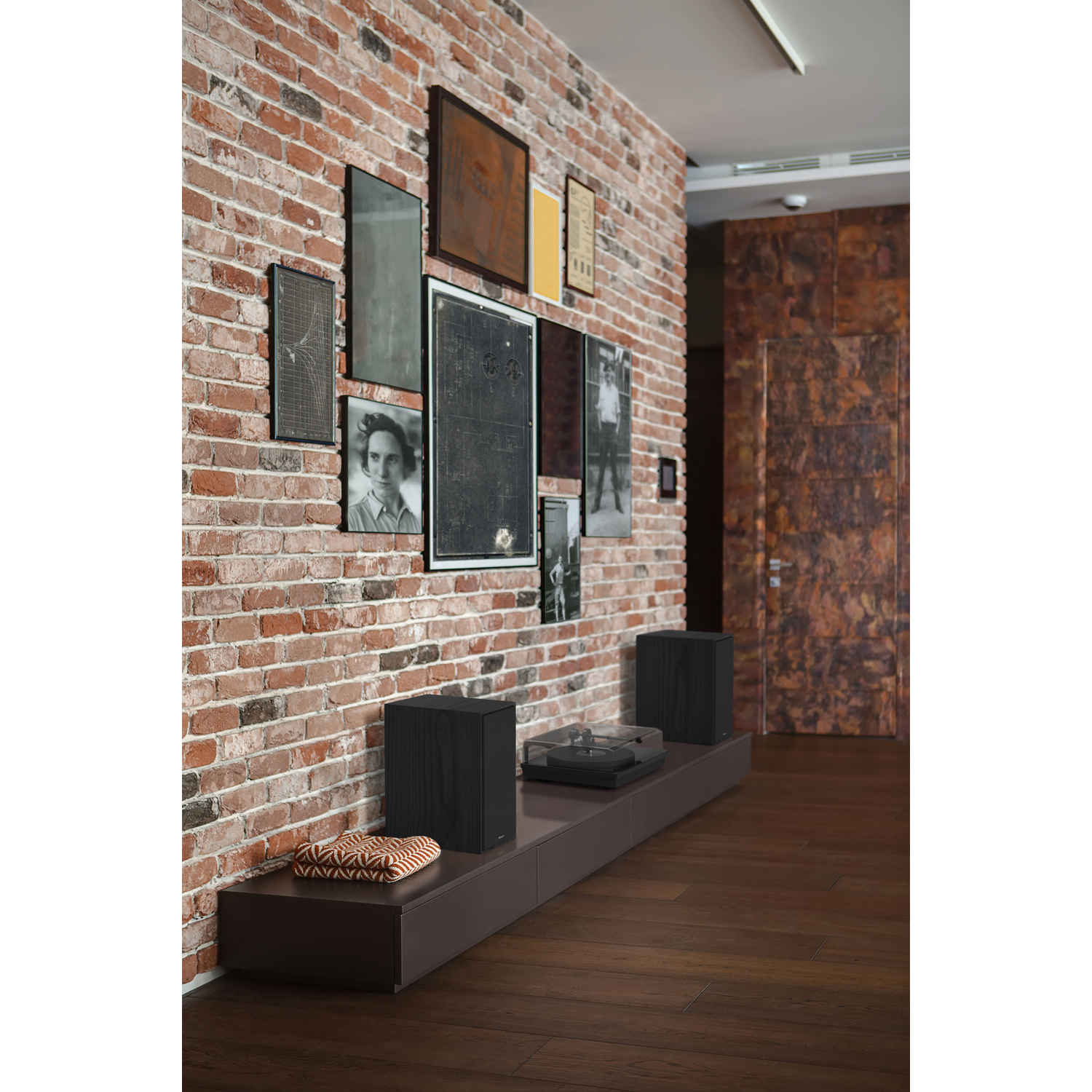 A room with speakers on a low cabinet, pictures on a brick wall, and a wood floor.