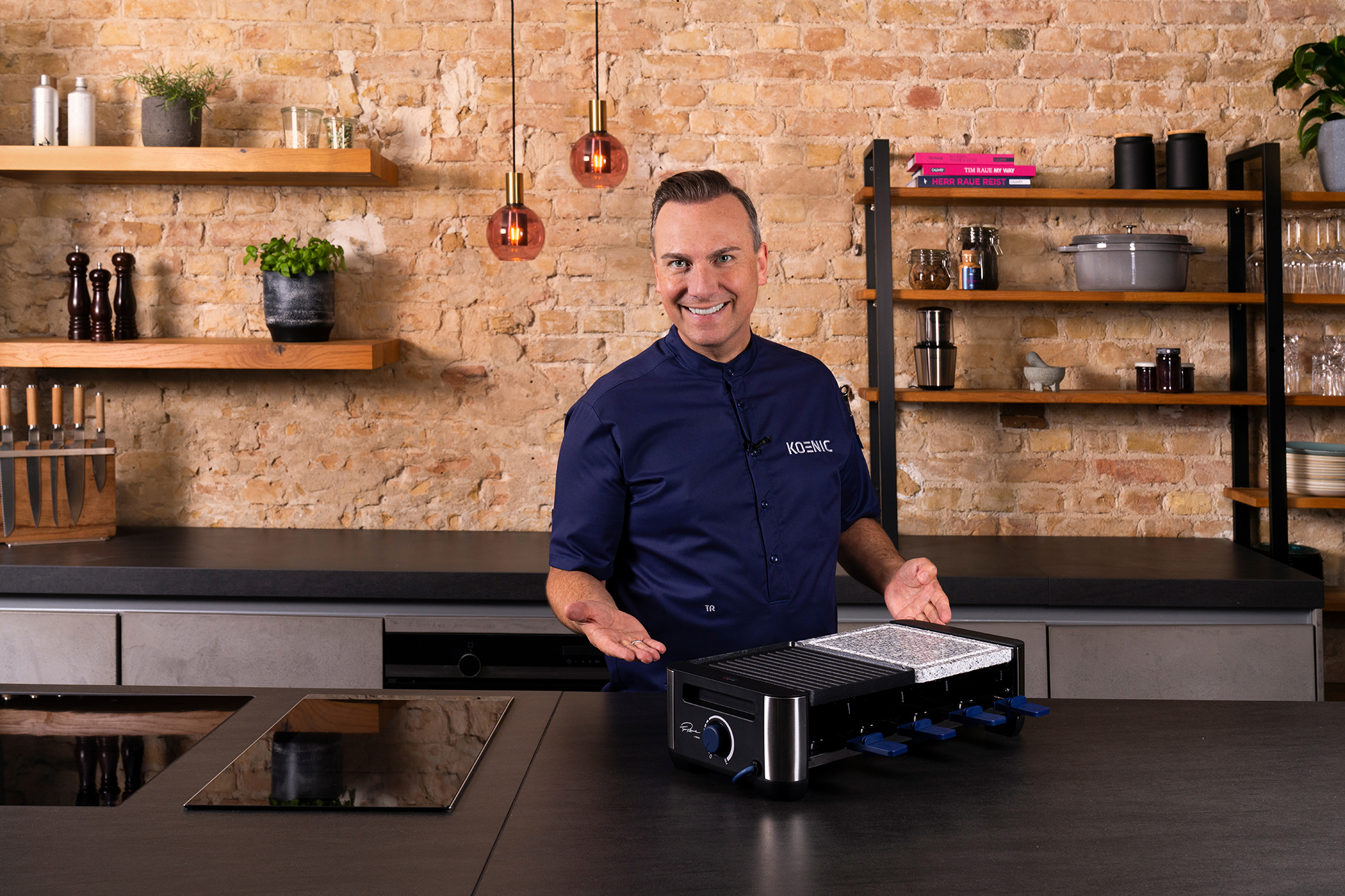 Ein Mann in einem Kochmantel lächelt und zeigt auf einen Raclette-Grill auf einer Küchentheke. Ziegelwand im Hintergrund.