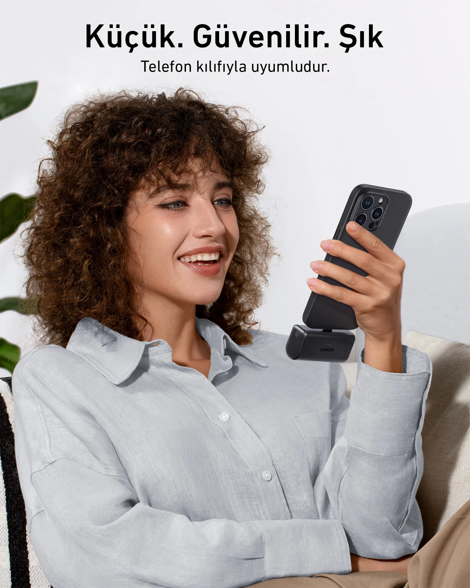 Gülümseyen kadın, küçük siyah bir şarj cihazına bağlı siyah bir telefon tutuyor. Bir kanepede oturuyor.