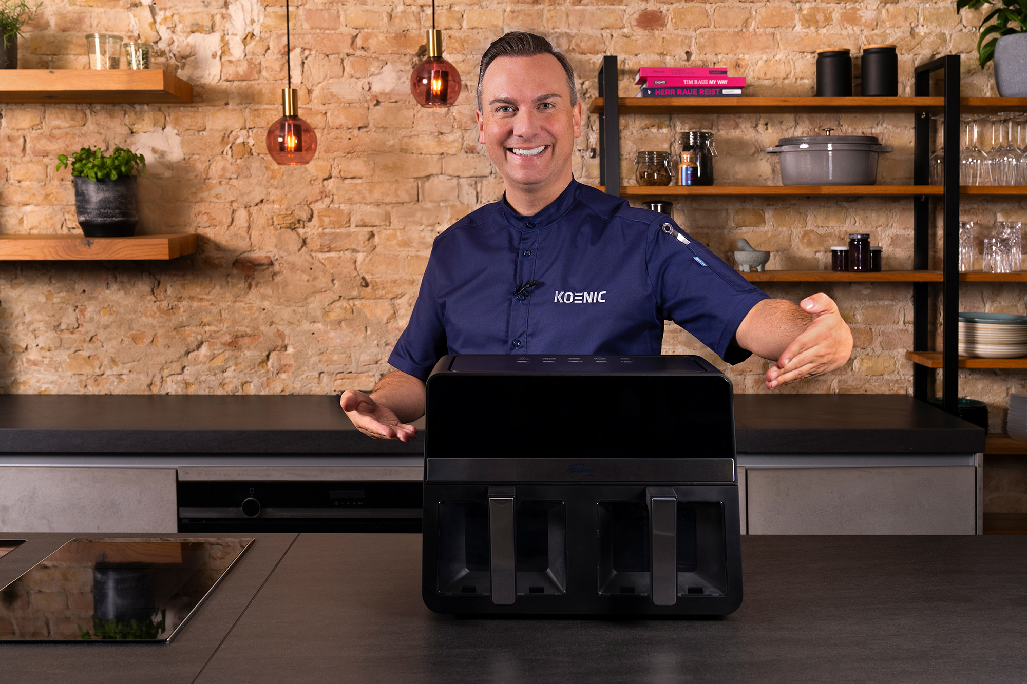 Ein Mann in einem dunkelblauen Kochmantel präsentiert eine schwarze Heißluftfritteuse. Küchenumgebung mit Ziegelmauer im Hintergrund.