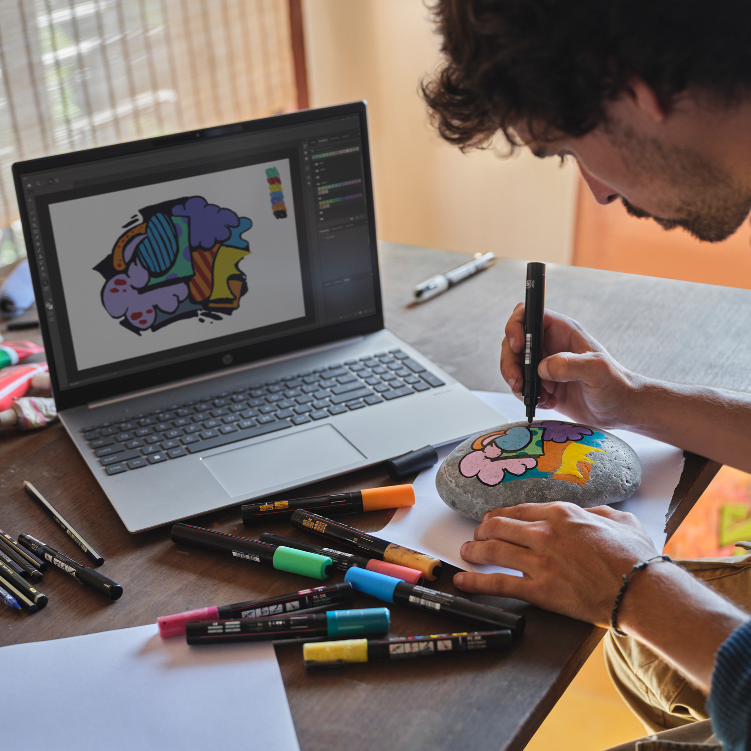 A man draws on a rock with colorful markers, using a laptop.