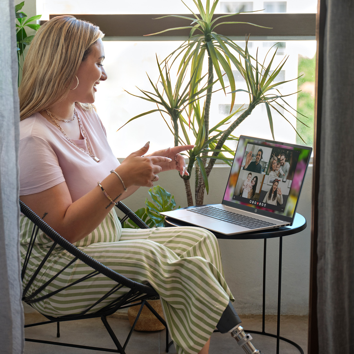 Woman in chair on balcony video calls on laptop. Plant in background.