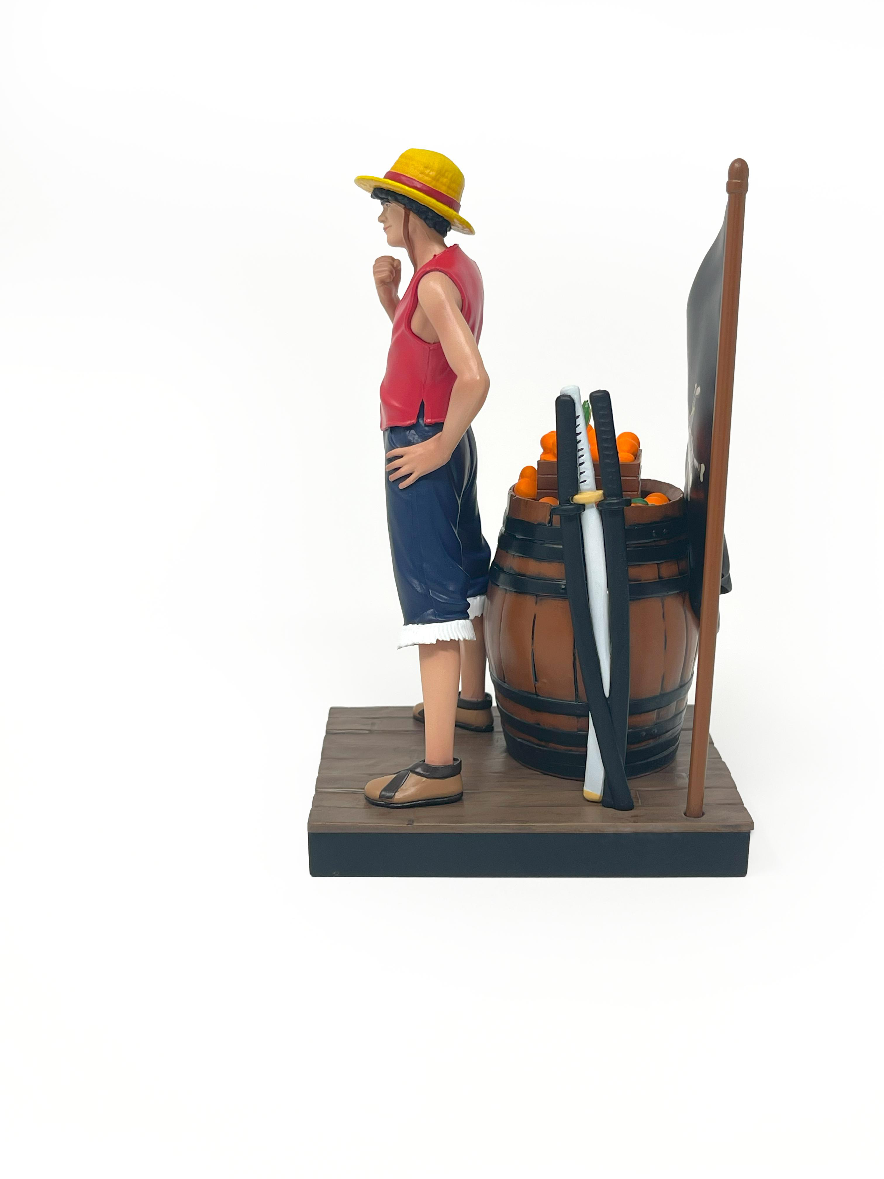 Luffy-Figur steht neben einem Fass voller Orangen, mit Schwertern und einer Flagge hinter ihm.