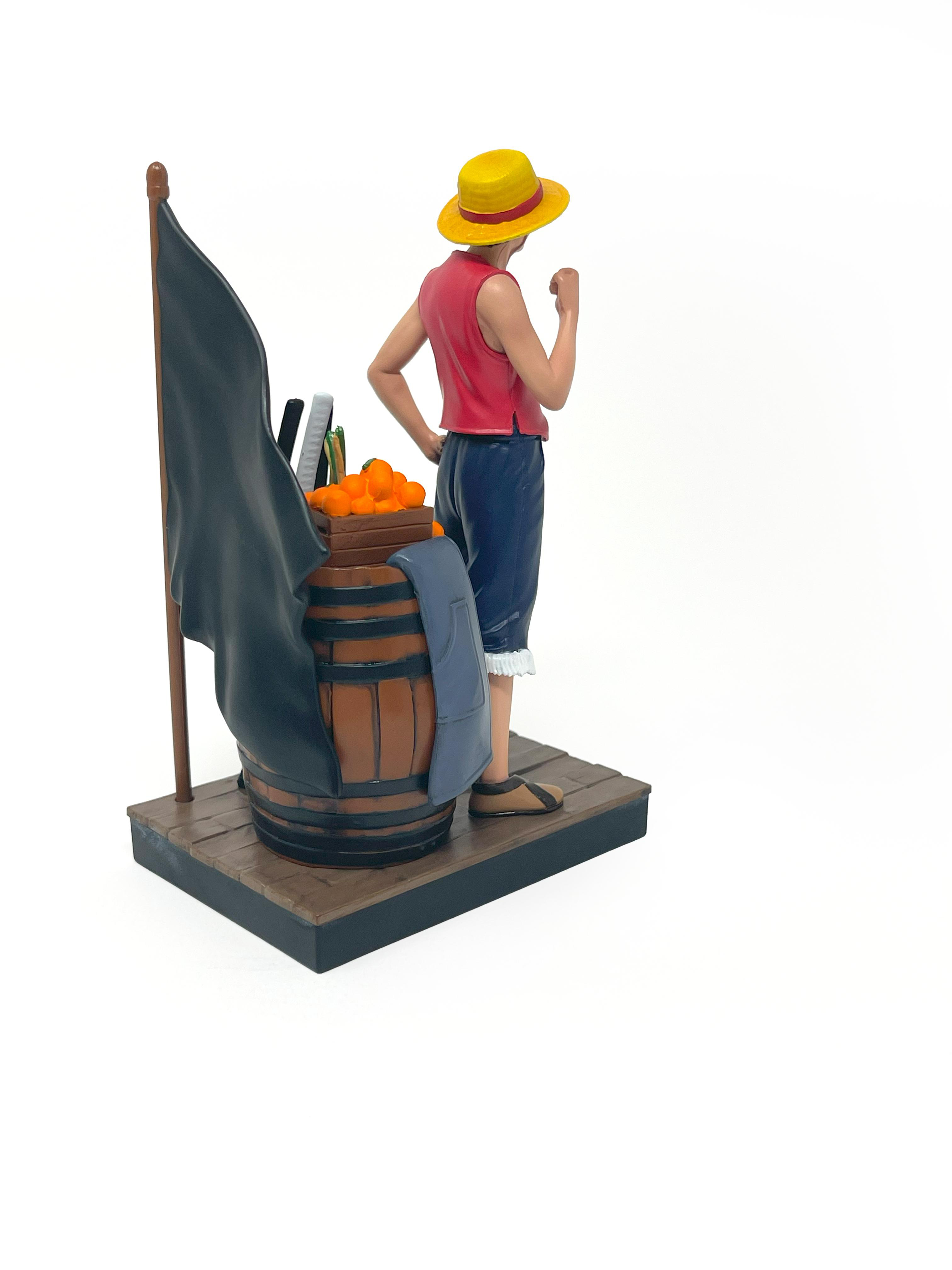 Luffy-Figur neben einem Fass mit Orangen, Schwertern, einer Flagge und einer Jacke. Weißer Hintergrund.