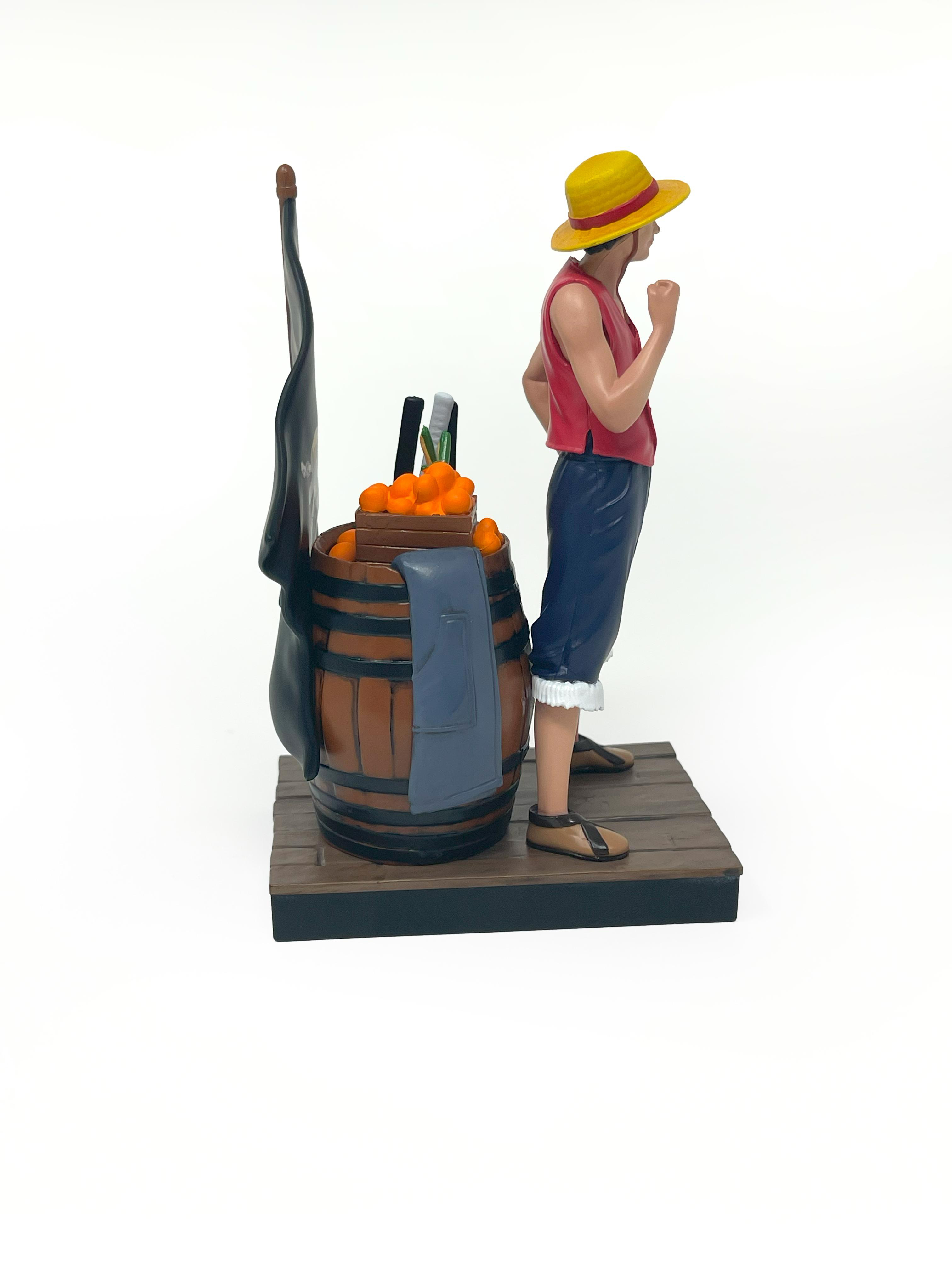 Luffy-Figur steht mit einem Fass voller Orangen, Schwertern, einer Flagge und einer Jacke. Weißer Hintergrund.