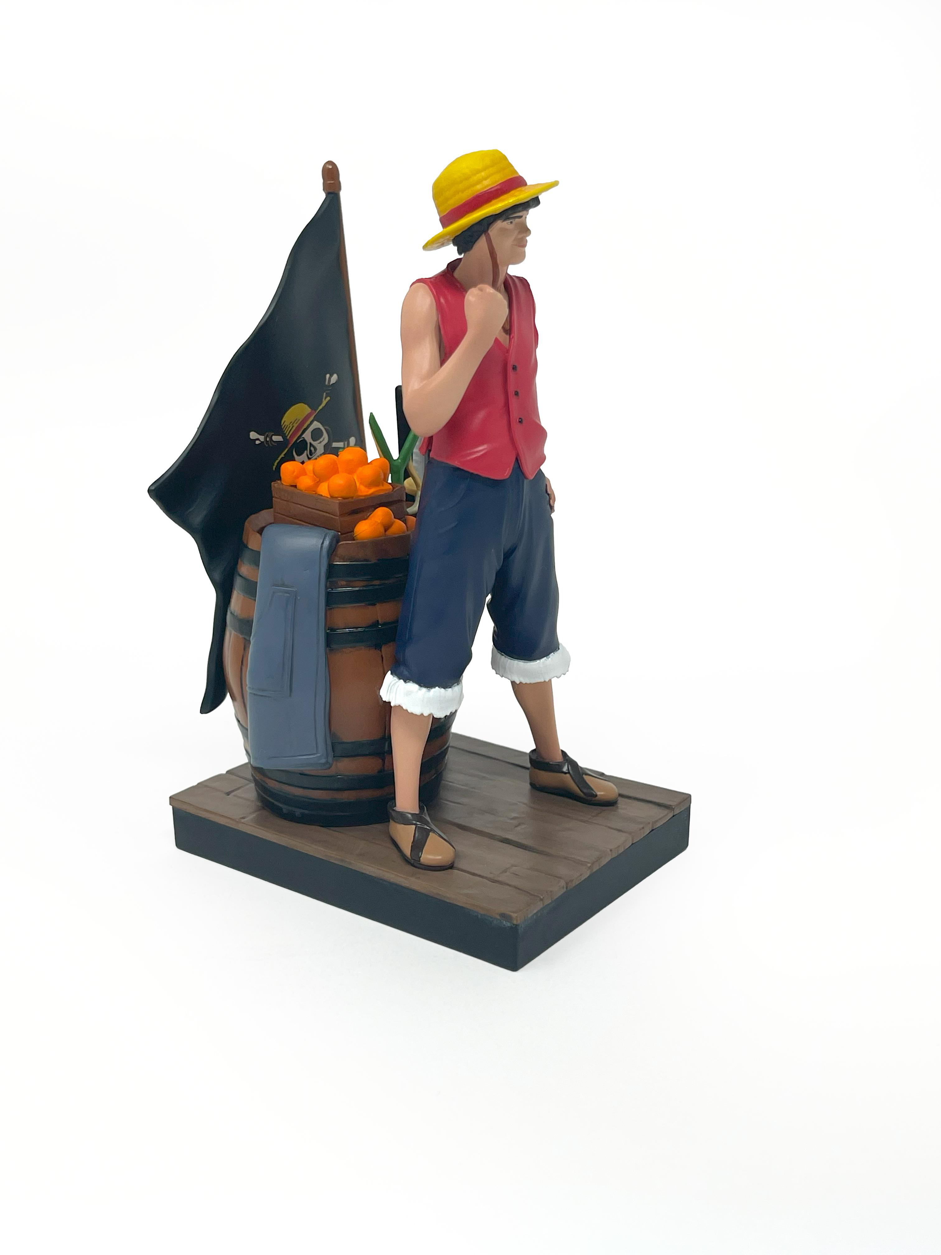 Luffy-Figur steht mit einem Fass, Orangen, einer Flagge und Schwertern. Weißer Hintergrund. Figur posiert.