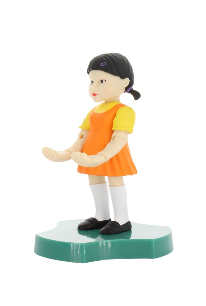 Kleine Puppe eines Mädchens in orangefarbenem Kleid mit gelben Ärmeln, weißen Socken und schwarzen Schuhen, stehend auf grünem Sockel.