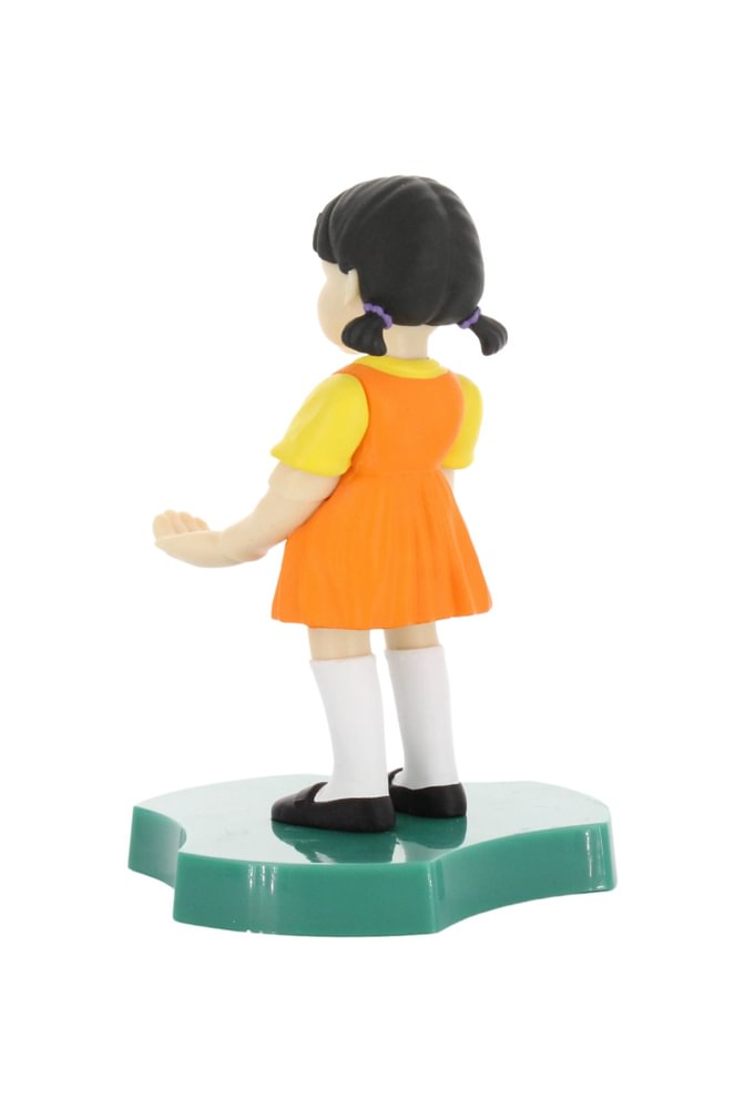 Die Puppe trägt ein orangefarbenes Kleid mit gelben Ärmeln, weiße Socken und schwarze Schuhe, steht auf grünem Sockel. Weißer Hintergrund.