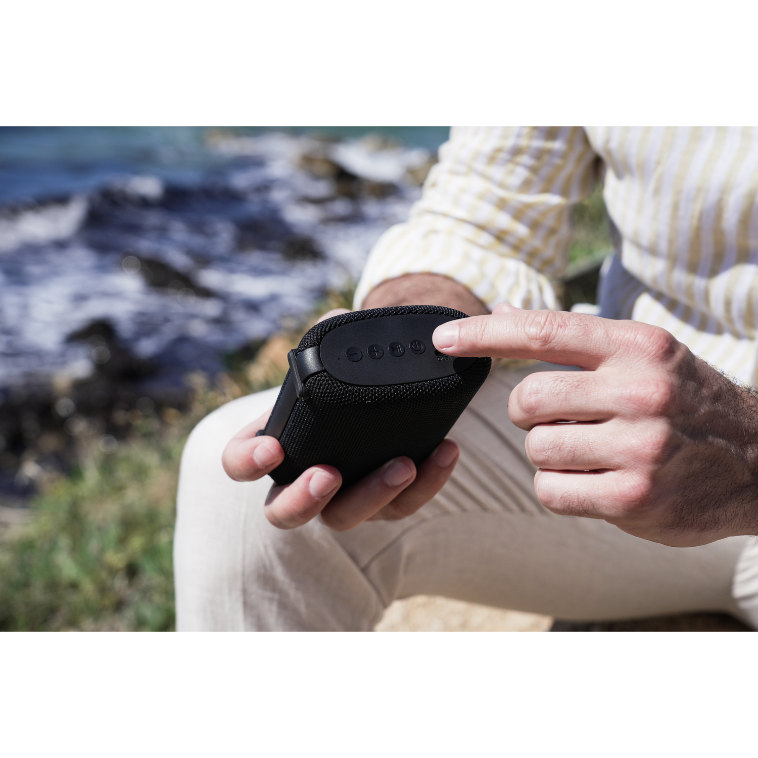 Man's hand touching speaker buttons. Black speaker. Background is ocean and sunny.