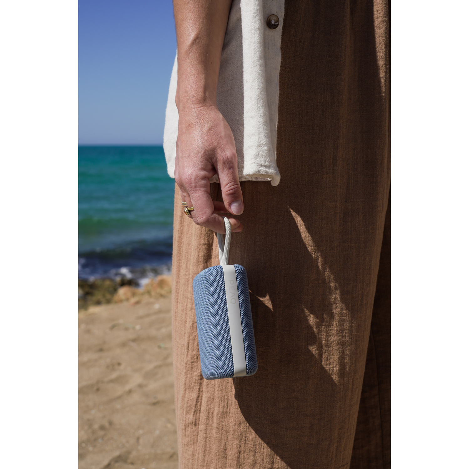 Hand houdt blauwe speaker vast. Strandachtergrond. Beige broek en wit shirt.