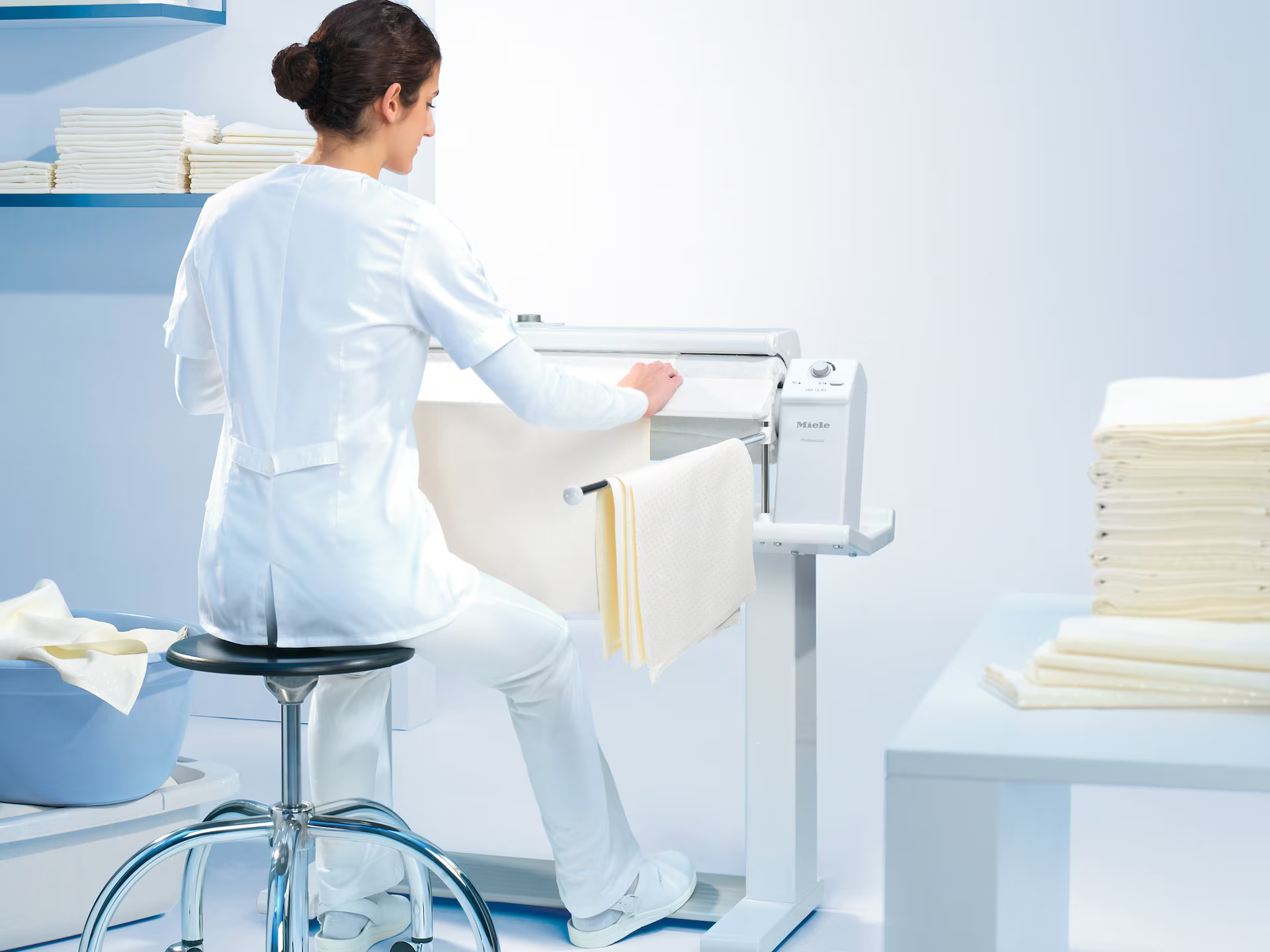 Eine Frau in weißer Uniform bügelt ein großes weißes Laken auf einer Bügelmaschine neben einem Stapel gefalteter Laken.