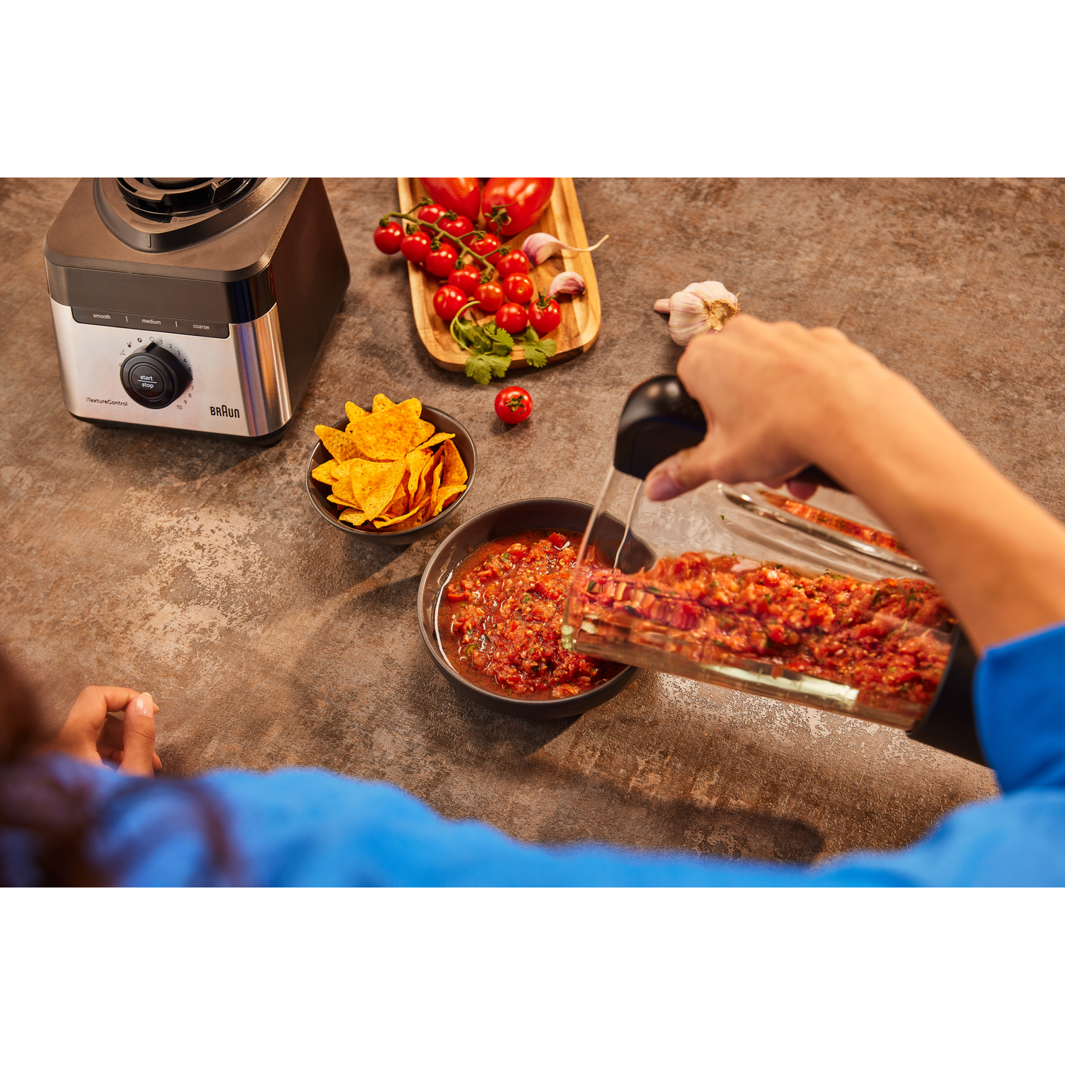 A person pouring salsa from a Braun blender into a bowl with chips and tomatoes.