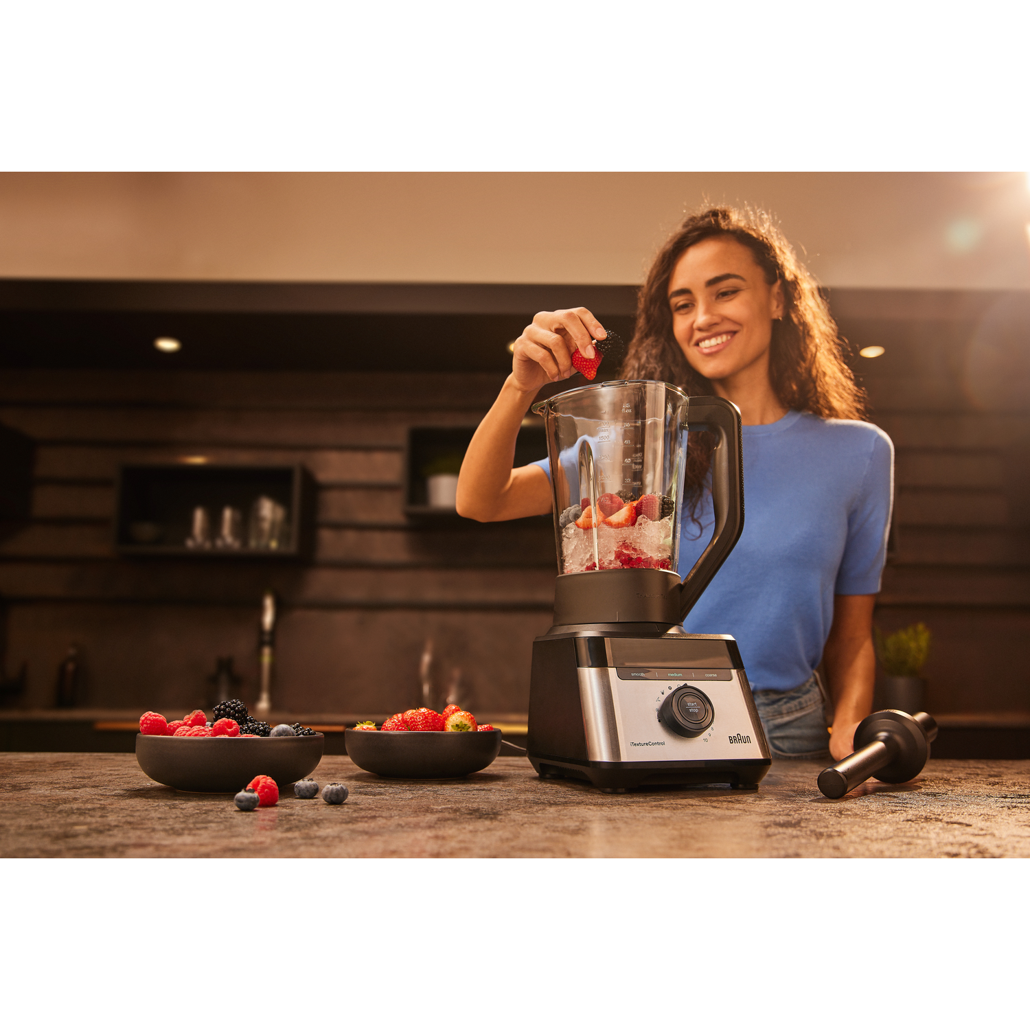 Woman adds strawberries to a blender with ice and other fruit.