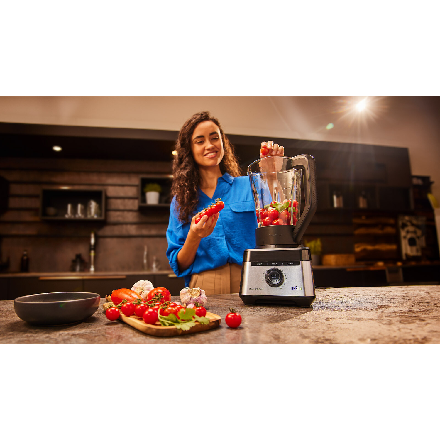 Woman is adding tomatoes to the blender in the kitchen.