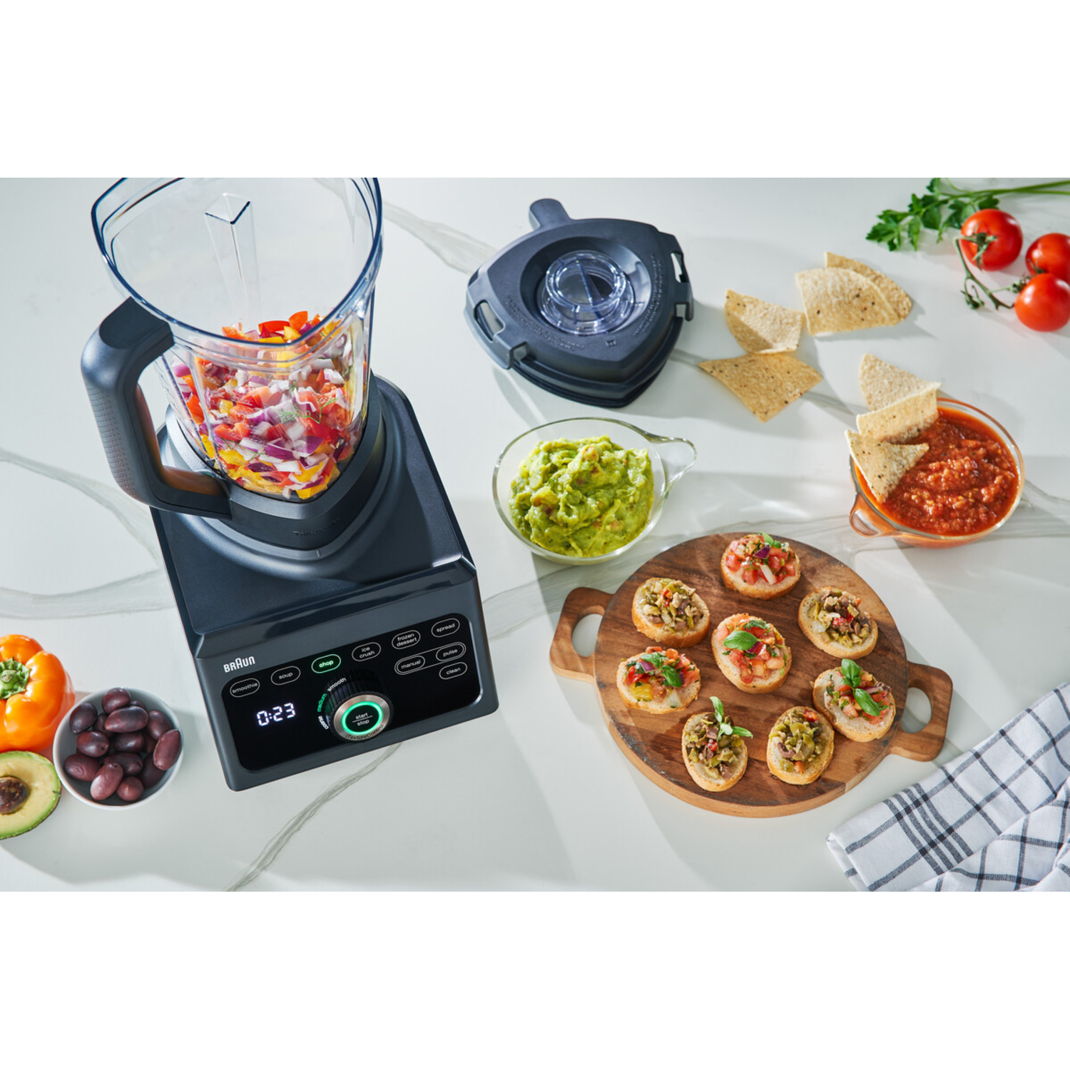 Blender with ingredients. Guacamole, salsa, and appetizers on a table. Tomatoes, avocados, and olives are present.