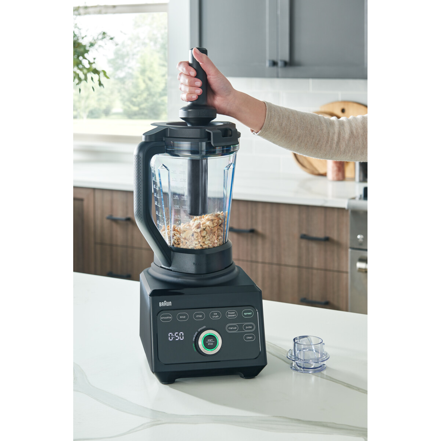 A person's hand using a black blender with a clear container, filled with nuts on a white surface.