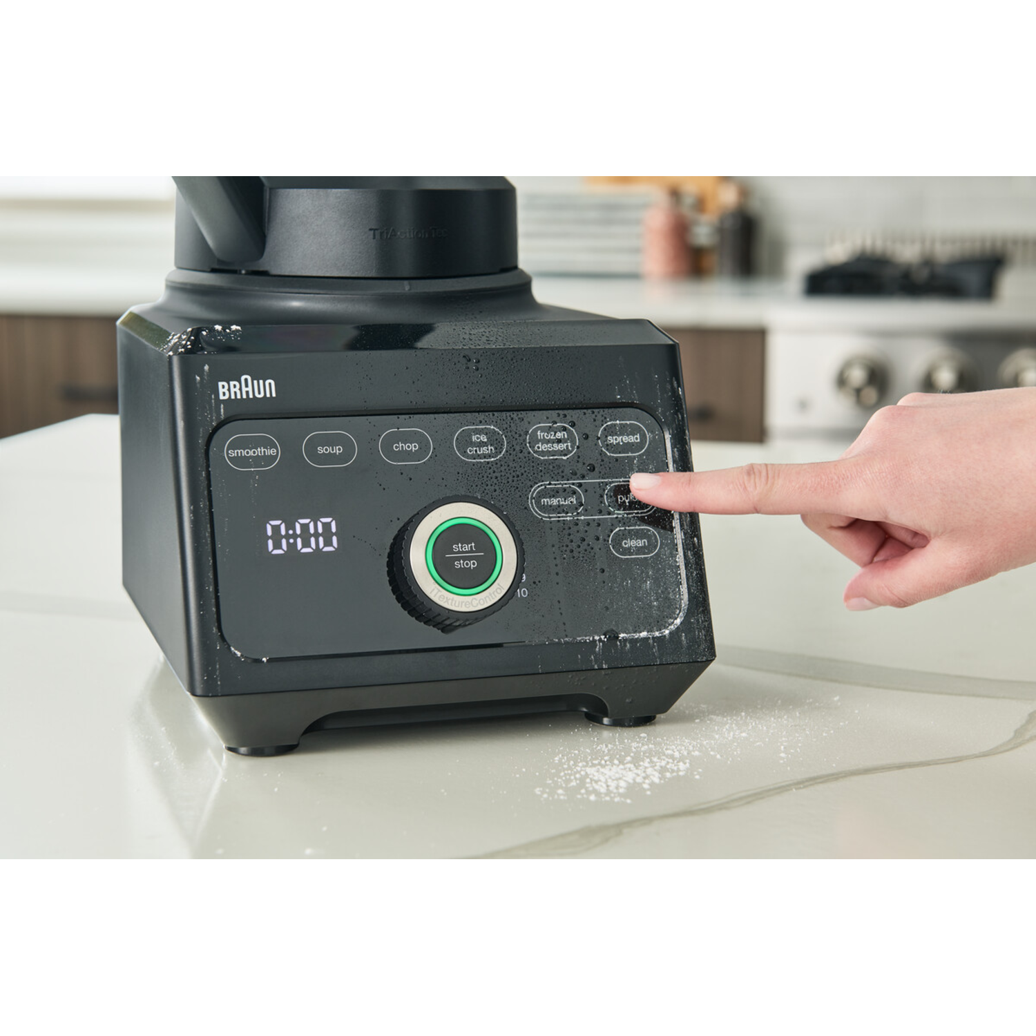 A black Braun blender. A finger presses a button. The display shows '0:00'. White countertop.