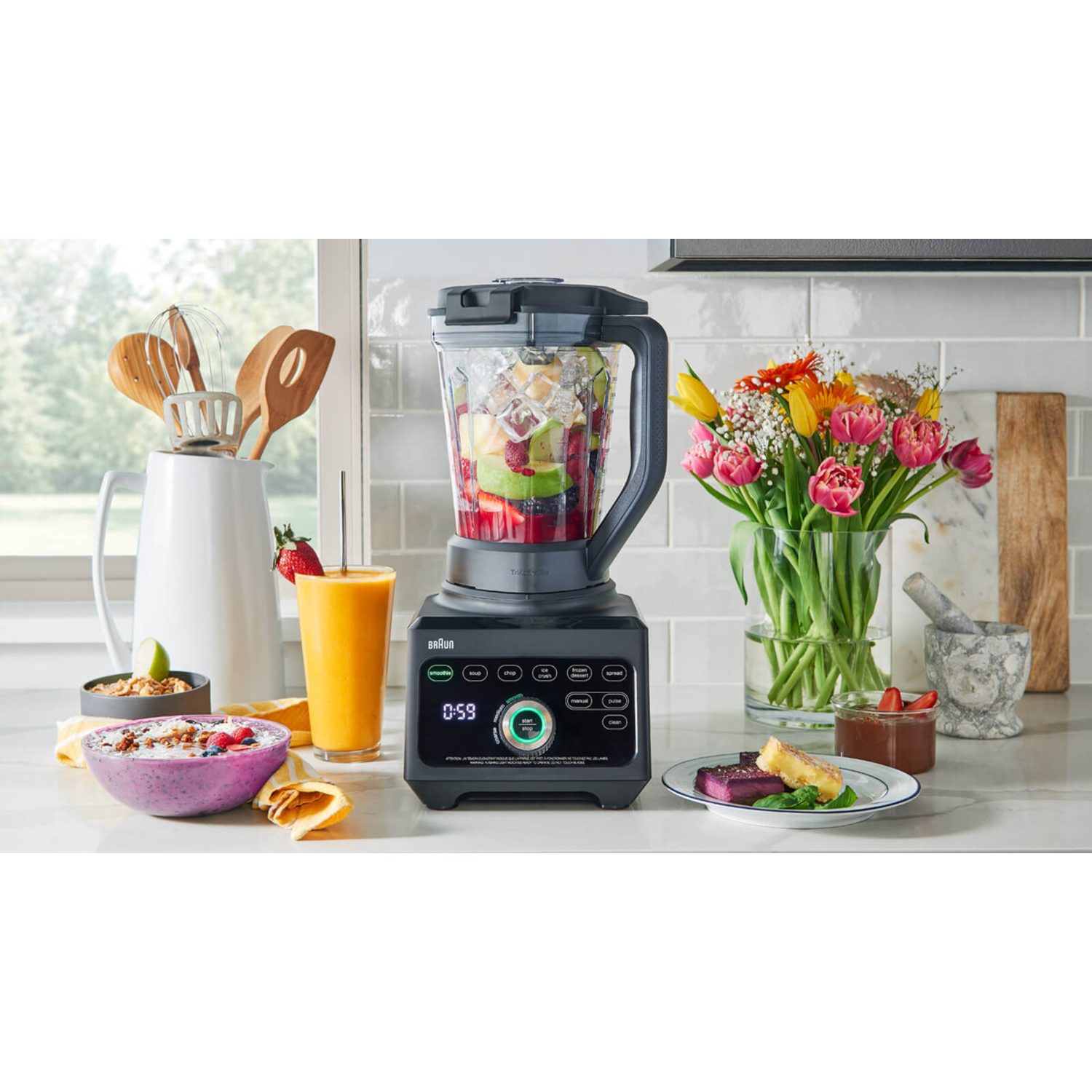 Braun blender with fruit and flowers. A glass of juice and food items are on the counter. Kitchen background.