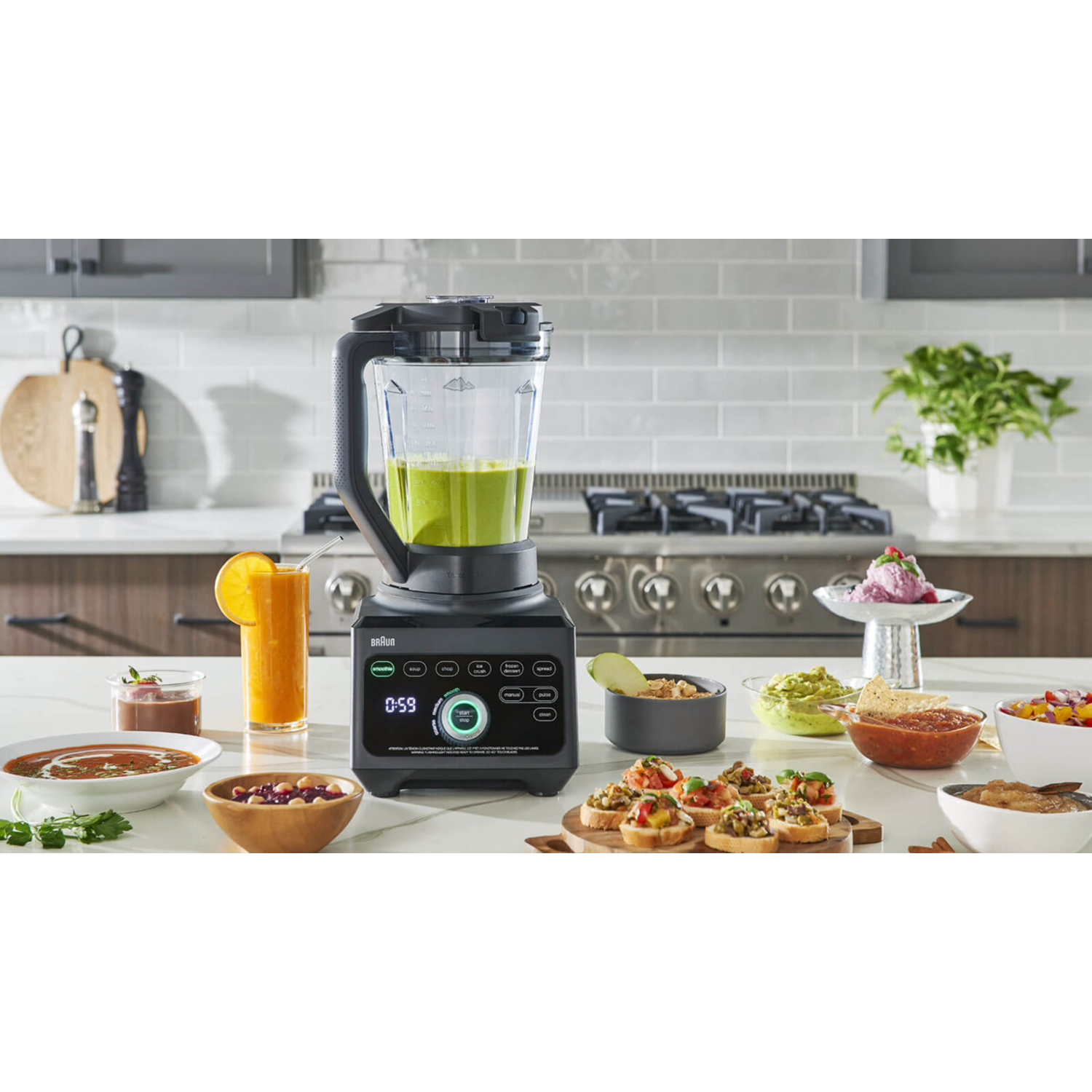 Braun blender with green mix. Various food items surround the blender. A kitchen setting.