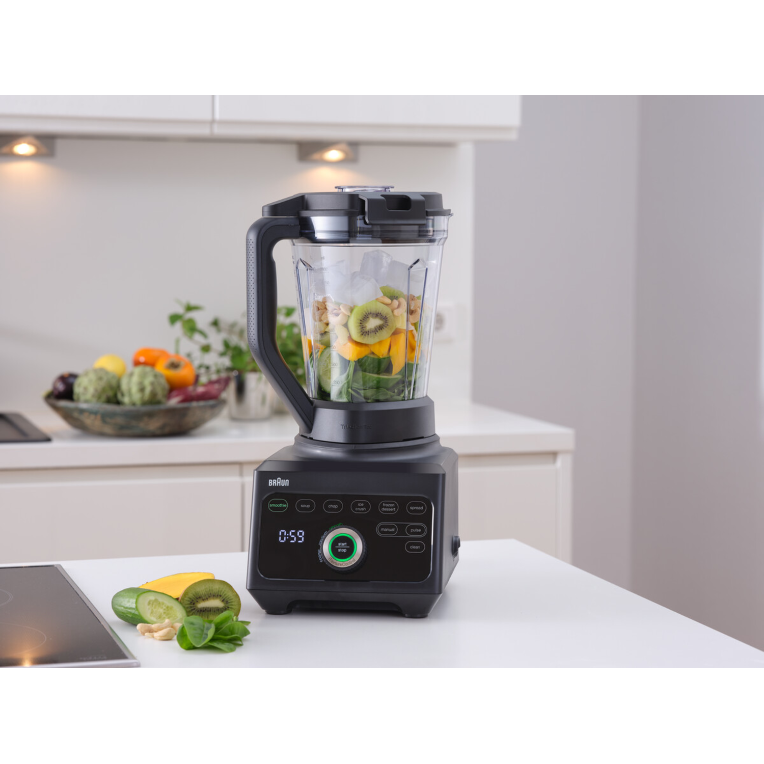 Braun blender with fruit inside. Kitchen counter with food items around. White cabinets in background.